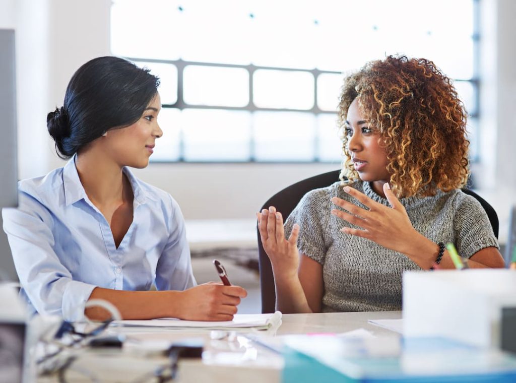 Manager and employee sitting at a table engaged in a conversation while taking notes on paper spread across the table