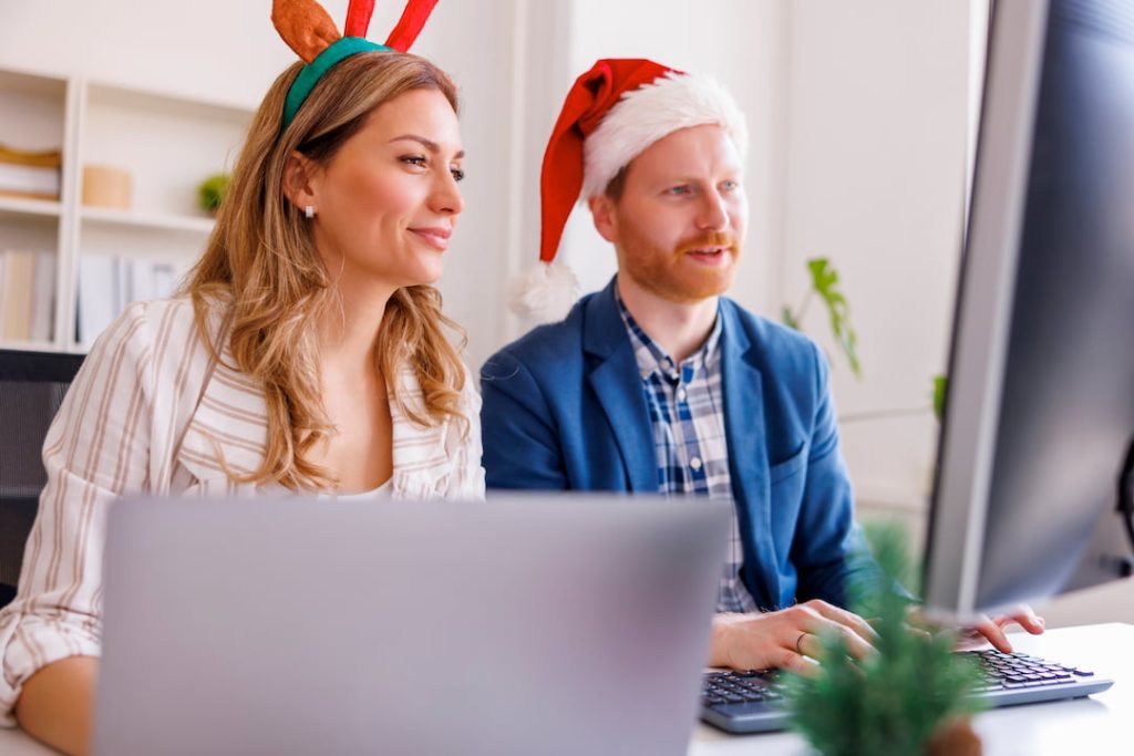 Two employees wearing festive holiday hats sitting at a desk in front of a laptop