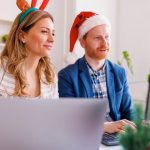 Two employees wearing festive holiday hats sitting at a desk in front of a laptop