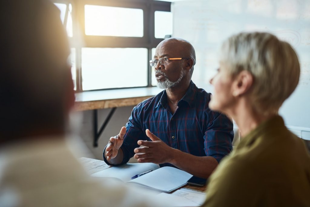 A business leader wearing glasses engaging in a conversation with coworkers around a table in an office building