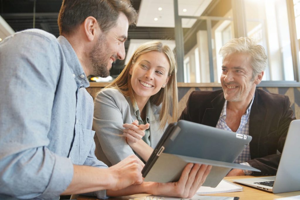 Group of three sales people around a table smiling, one of whom is showing the other two something on an ipad.