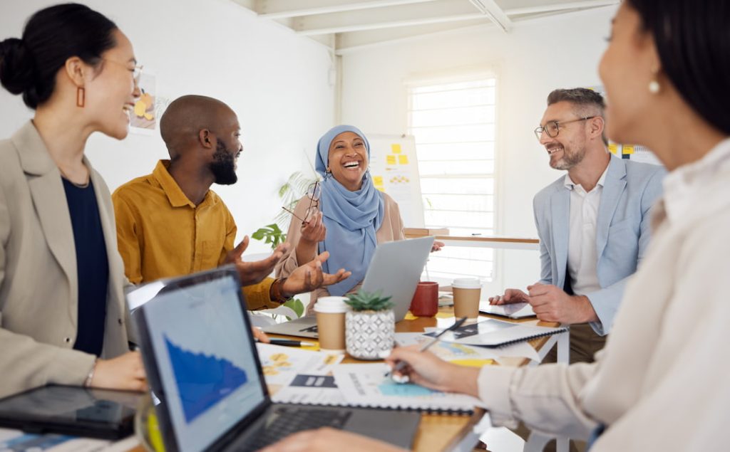 Diverse group of employees sitting around a table and laughing together while working on their laptops